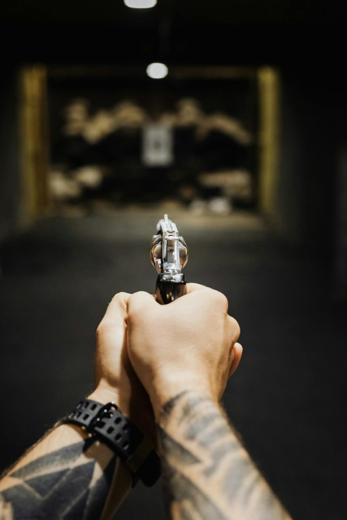 Hands with tattoos aiming a revolver in an indoor shooting range, focused on gun detail.